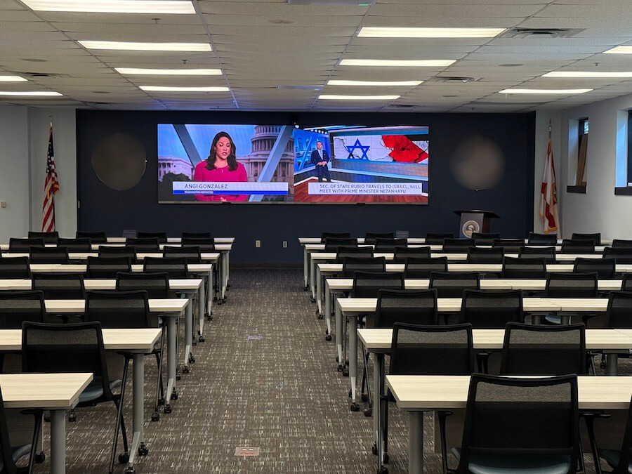 A classroom with rows of tables and desks facing a split screen. One side of the screen shows a news caster and the other shows news about Israel.
