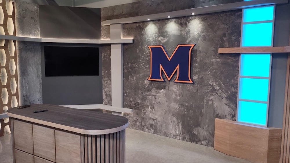 Broadcast-style studio set with illuminated anchor desk, accent lighting, wall-mounted display, and professional set design