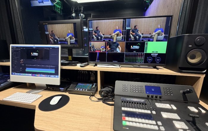 Broadcast control room with multi-view monitors, custom shallow console, and professional audio mixing equipment.